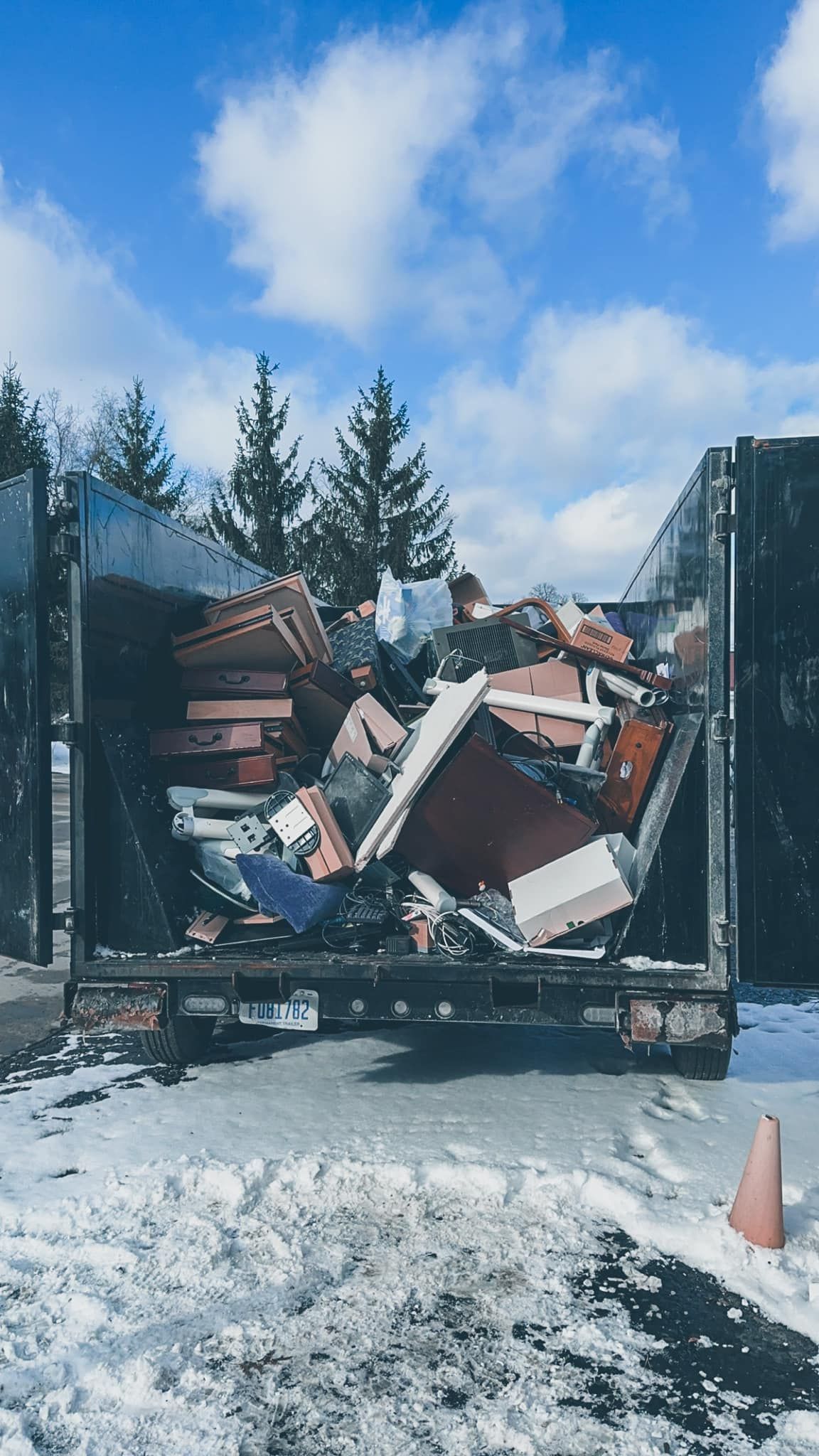 Trash-filled dumpster truck in snowy lot, with pine trees and blue sky in the background.