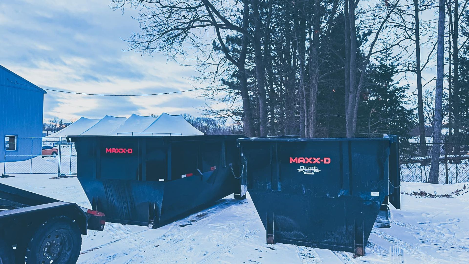 Two black dumpsters on a snowy street beside a blue building and tall trees