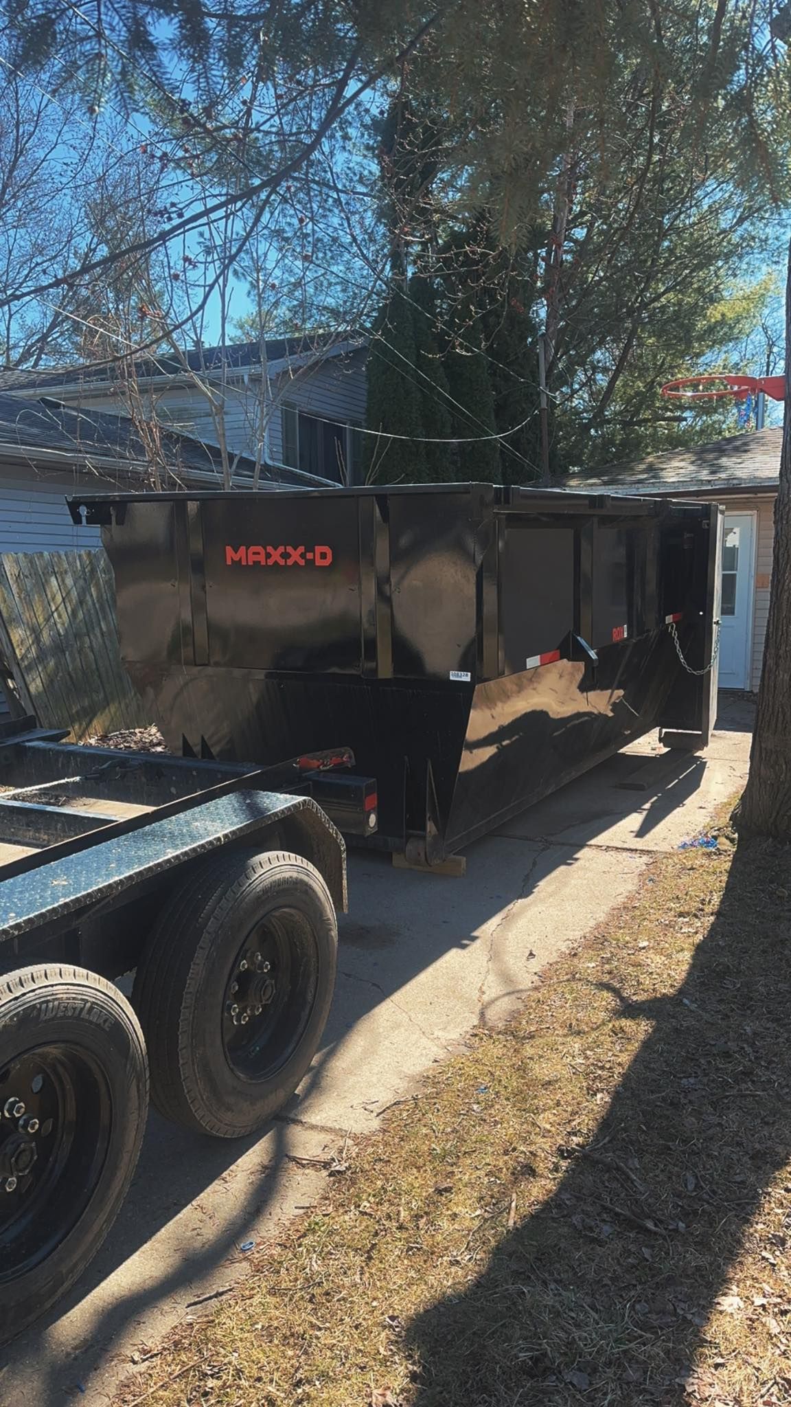 Black dump trailer parked in a driveway beside a house, with trees overhead and sunlit ground.
