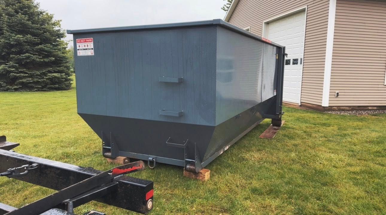 Blue metal dumpster trailer beside a beige house on a grassy yard, with a black hitch in front.