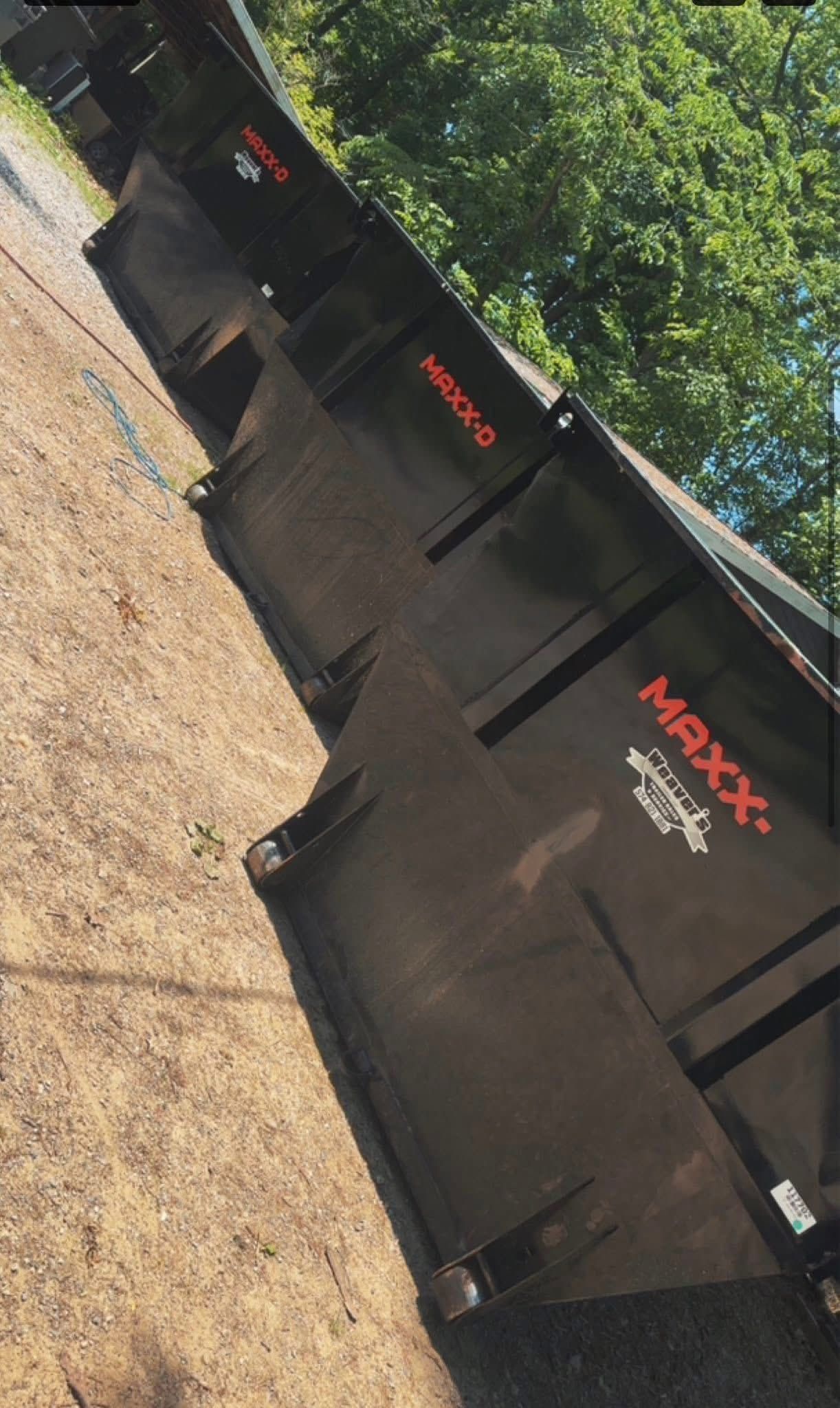 Black Maxx dumpsters lined up along a gravel lot beside trees, viewed at an angle.
