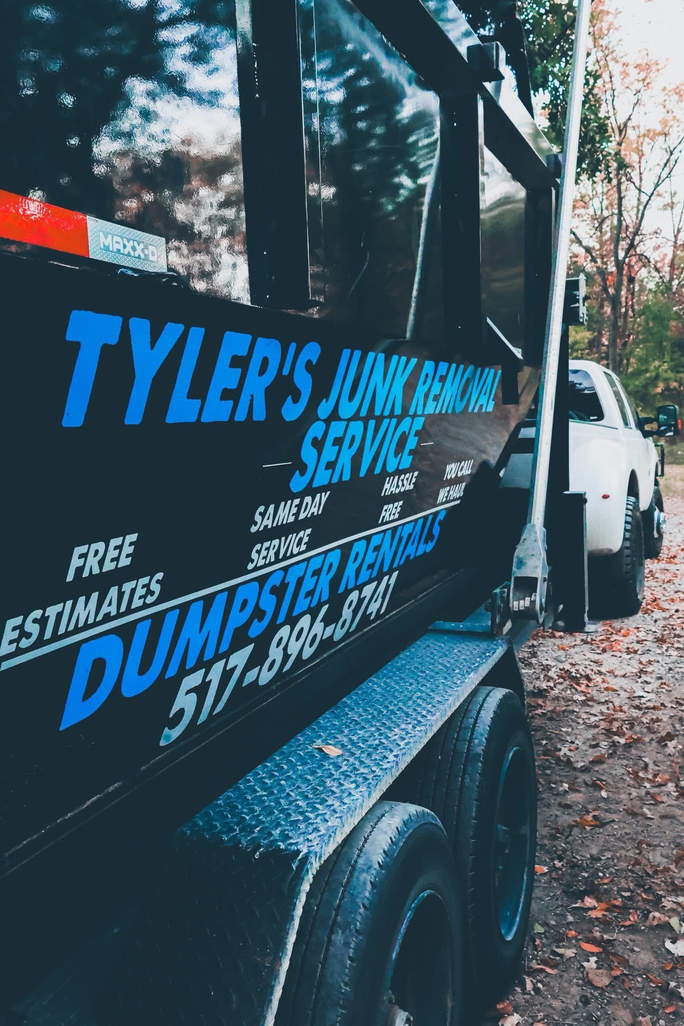 Blue dumpster trailer with Tyler’s Dumpster Service lettering parked beside a white truck on a tree-lined street