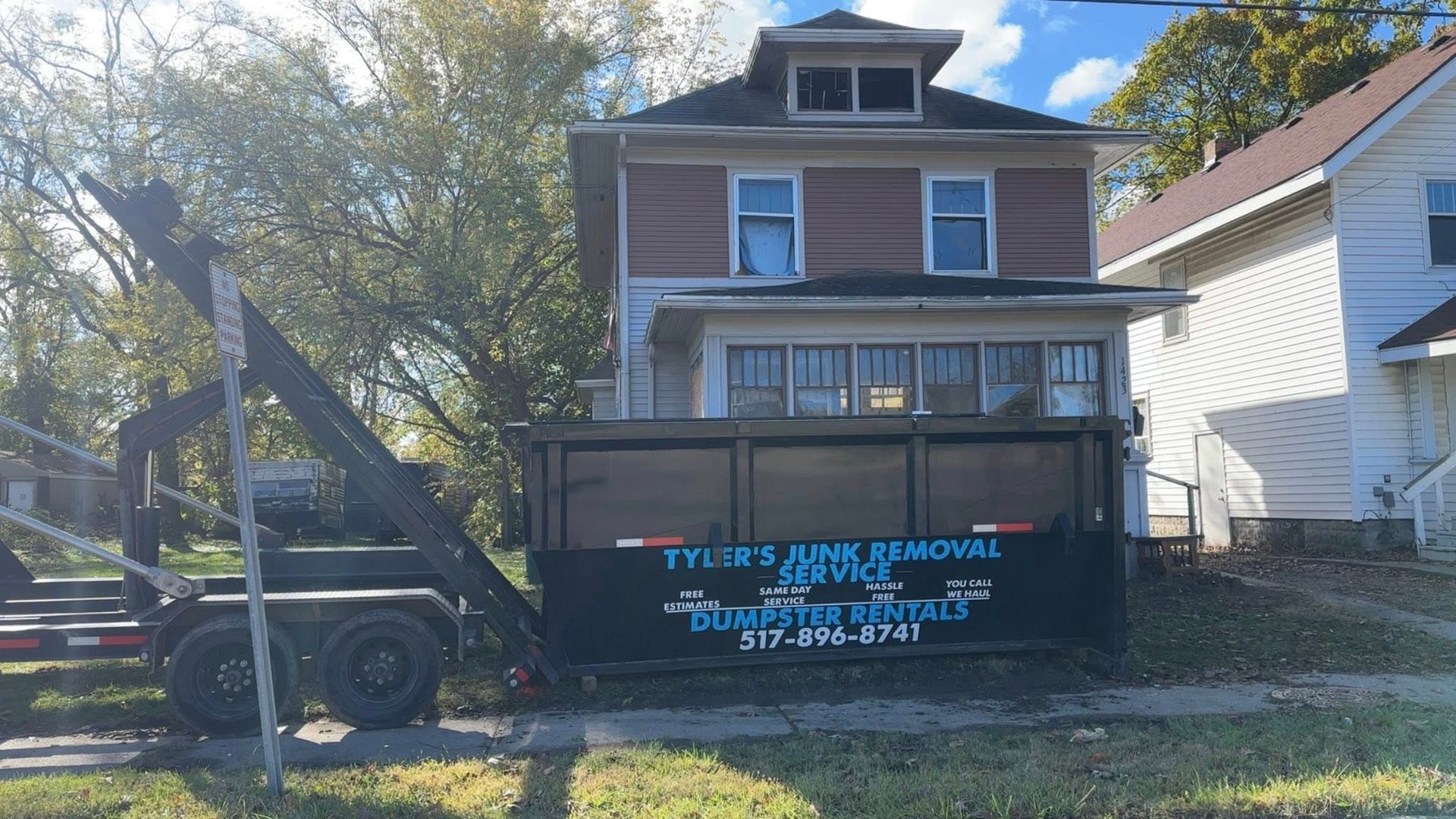 A blue dumpster in front of a three-story house, with a tow truck parked on the left.