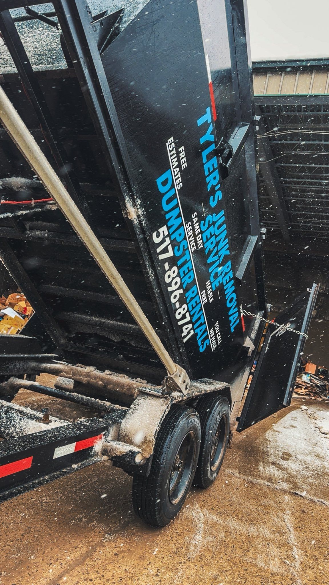 Black trailer with blue and white lettering, parked on wet ground near a building.