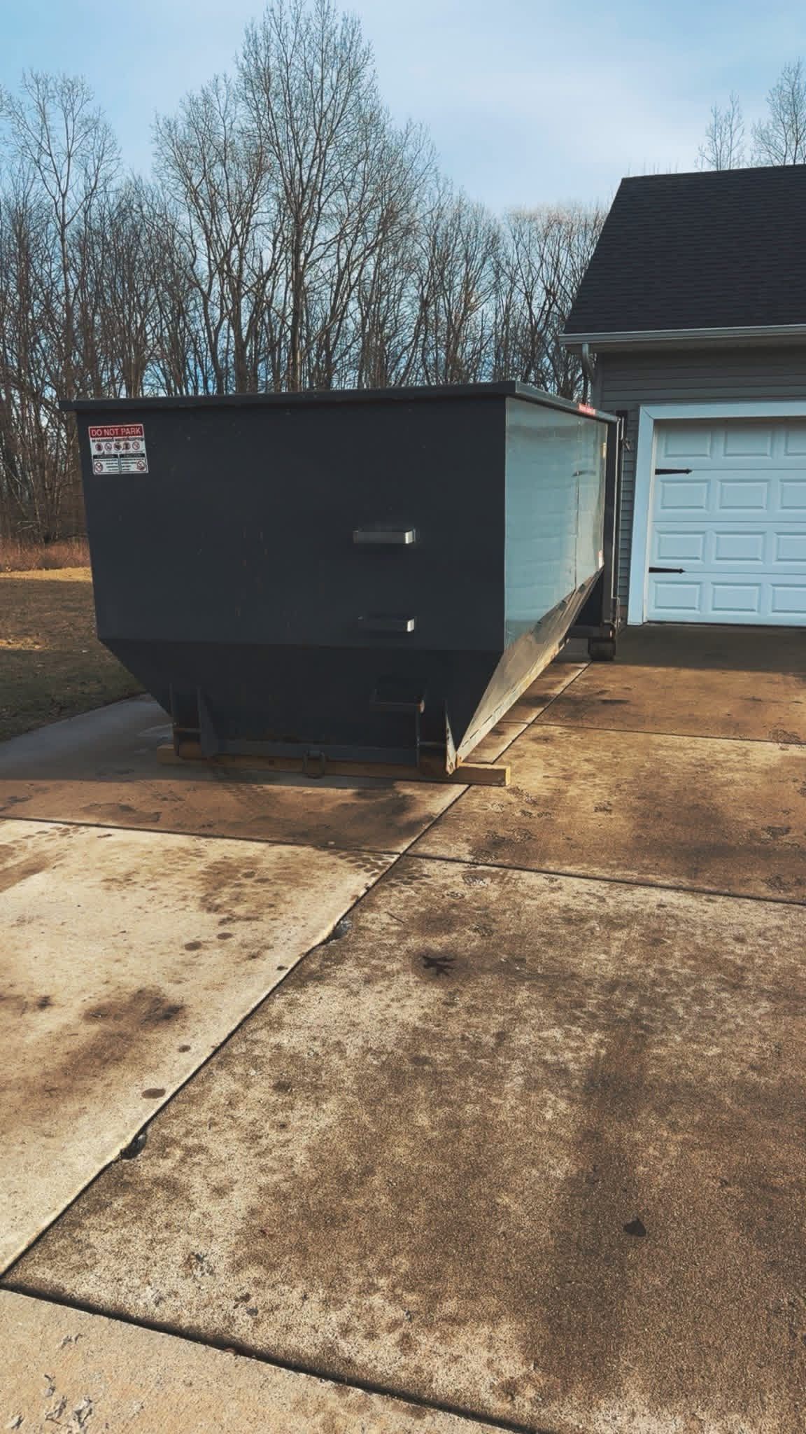 Black dumpster beside a garage on a driveway, with trees in the background.