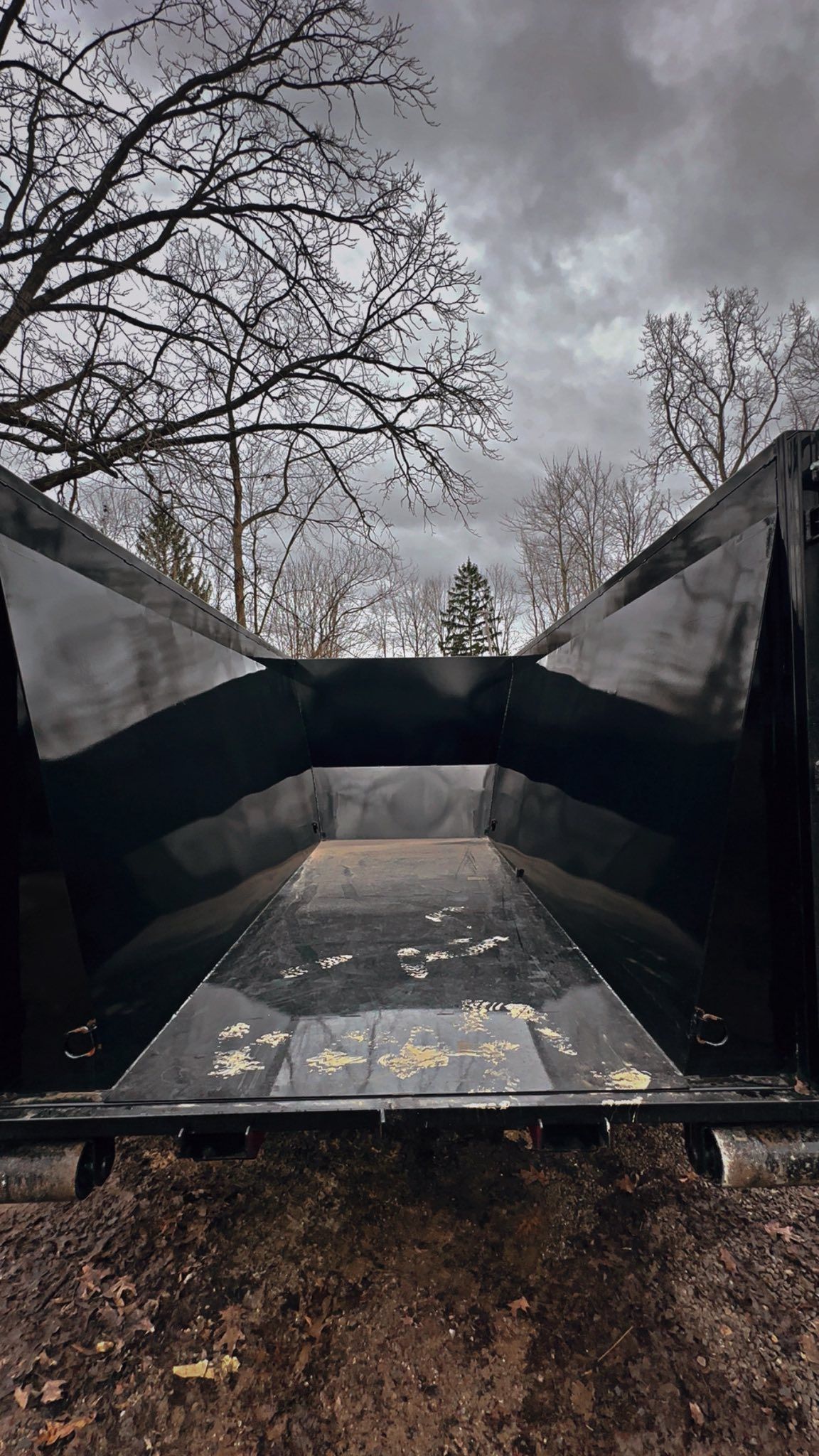 Black metal dumpster or container in a wooded outdoor area under a cloudy sky