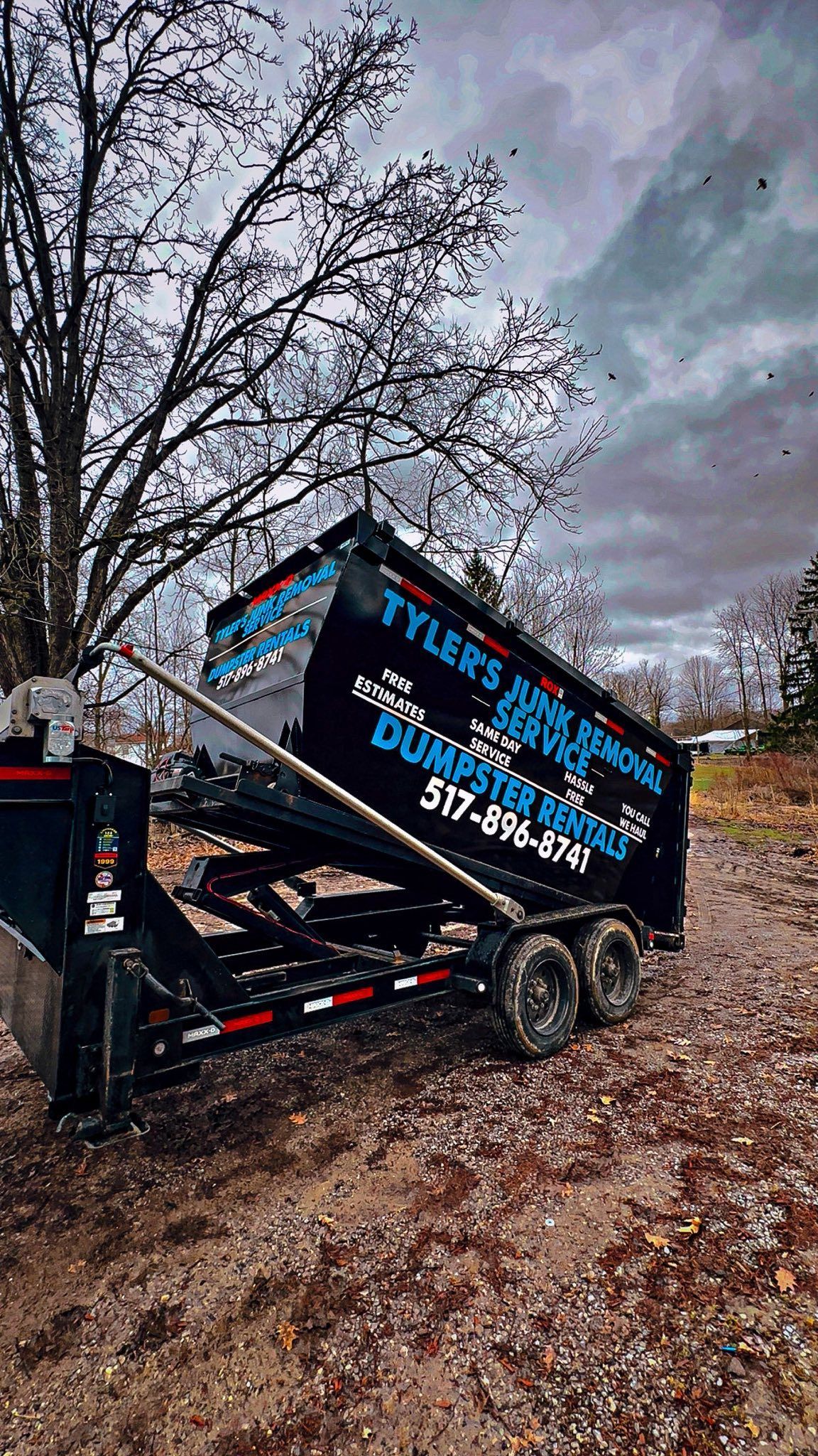 Black landscaping trailer with blue lettering parked on a leaf-covered roadside under cloudy sky