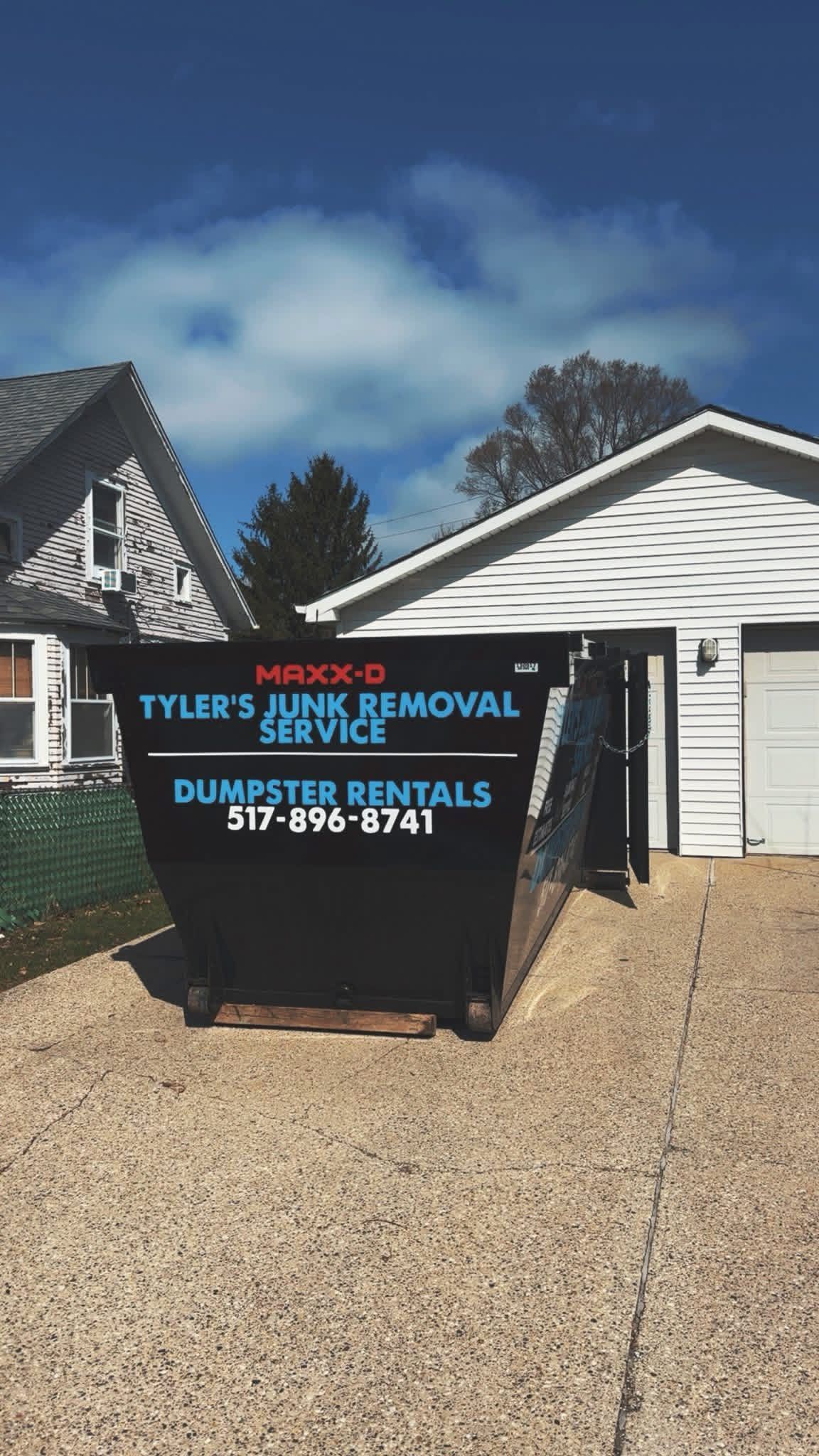 Black dumpster in a gravel driveway beside a house, with turquoise and orange service lettering.
