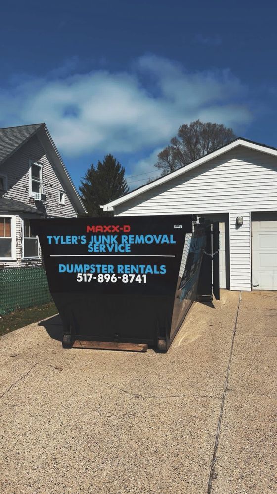 Black dumpster in a gravel driveway beside a house, with turquoise and orange service lettering.