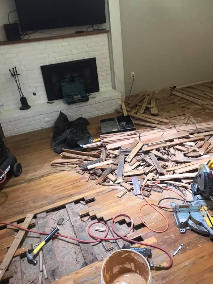 Living room undergoing flooring demolition; debris, tools, fireplace, and exposed subfloor are visible.