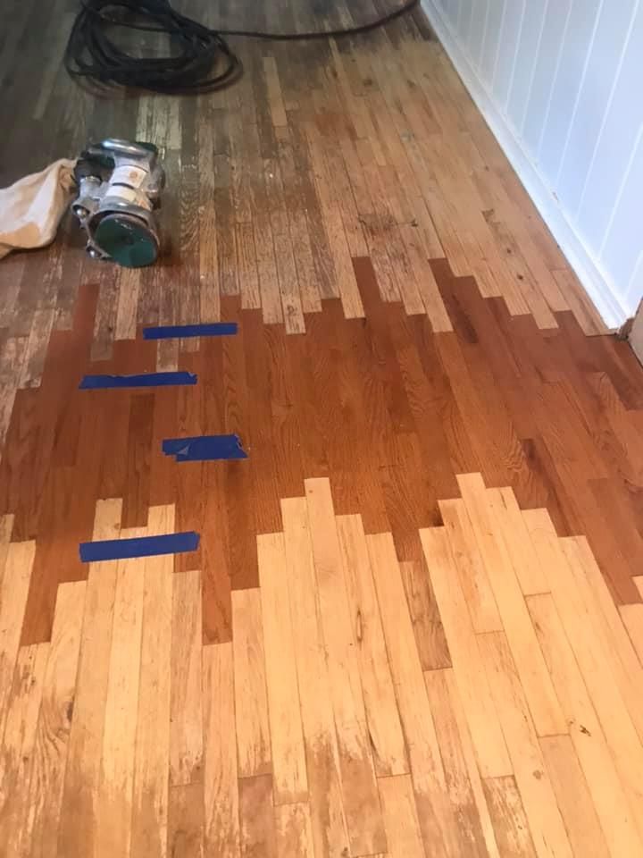 Hardwood floor being refinished, showing sanded wood next to stained section with blue tape.