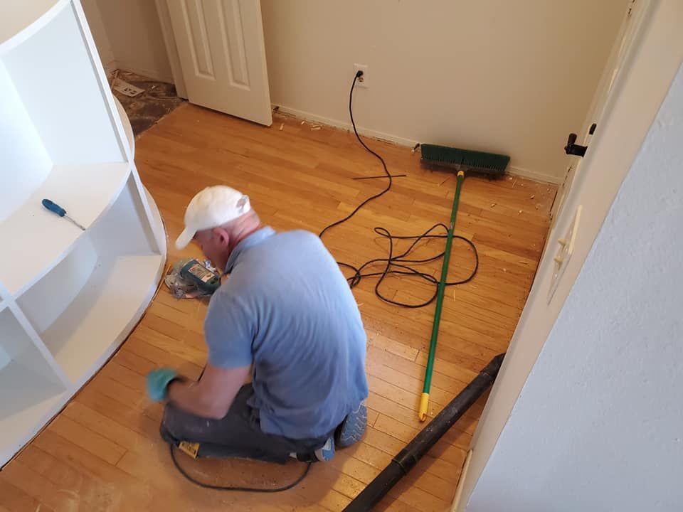 Person sanding a hardwood floor in a room. White shelves, a door, and vacuum are visible.