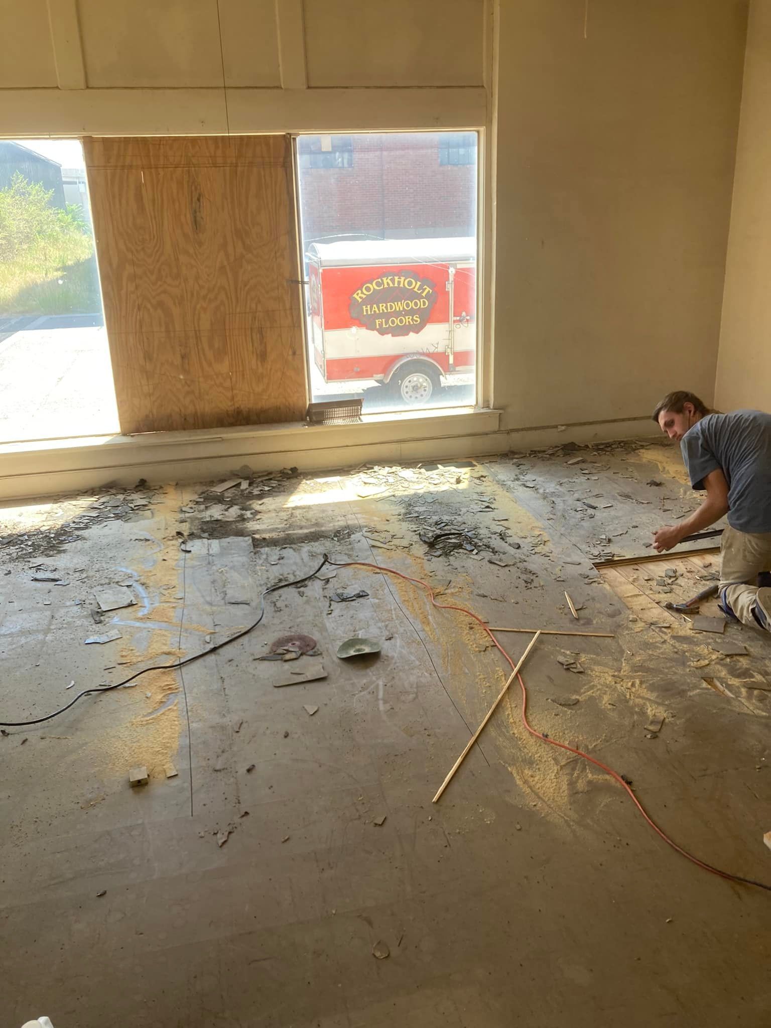 Person kneeling, examining debris on a concrete floor near a boarded-up window with a firetruck visible outside.