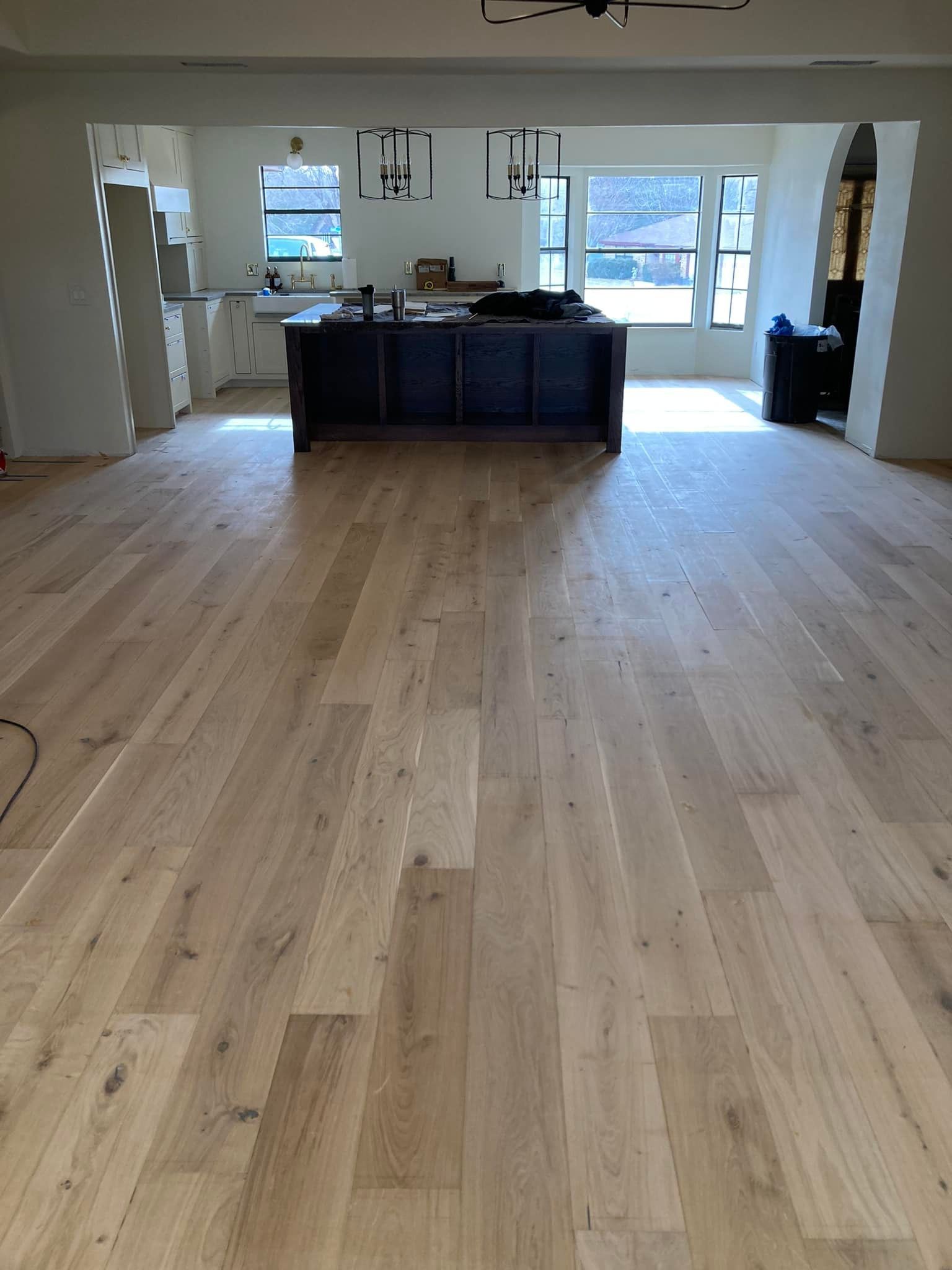 Light-colored wood flooring in a large room, leading to a dark kitchen island. White walls and windows are visible.