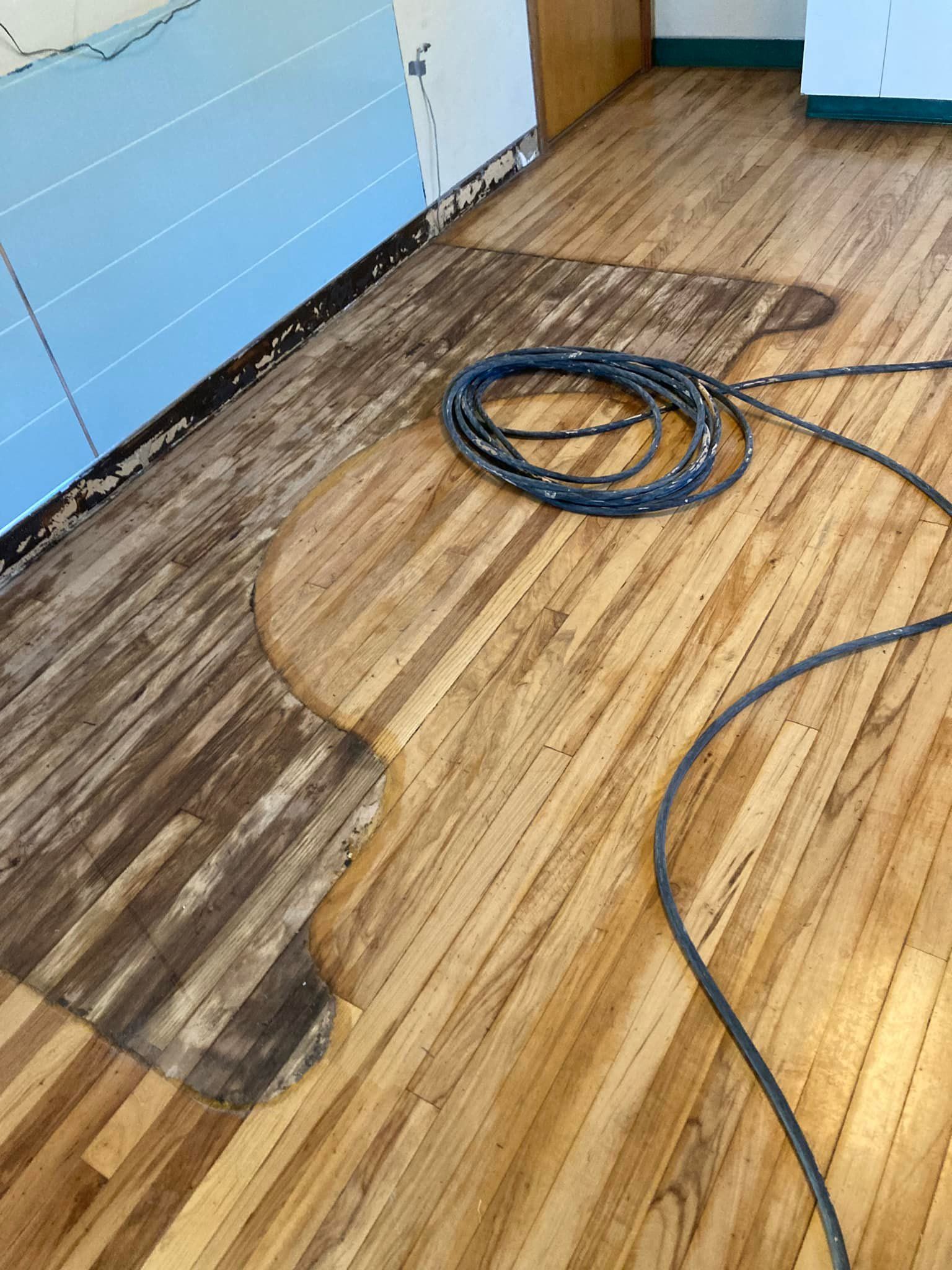 Wood floor being refinished; one section sanded, showing lighter wood, rest in darker state with cord.