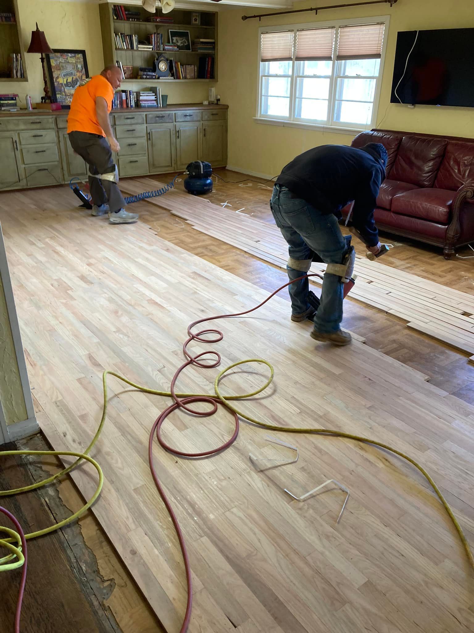 Two workers installing hardwood floor in a room with a red leather couch and cabinets.