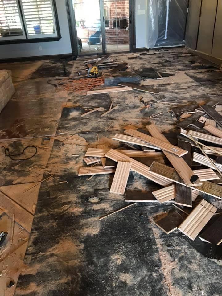 Debris from flooring removal. Scattered wood pieces, dust, and tools on a dark surface in a room with windows and a door.