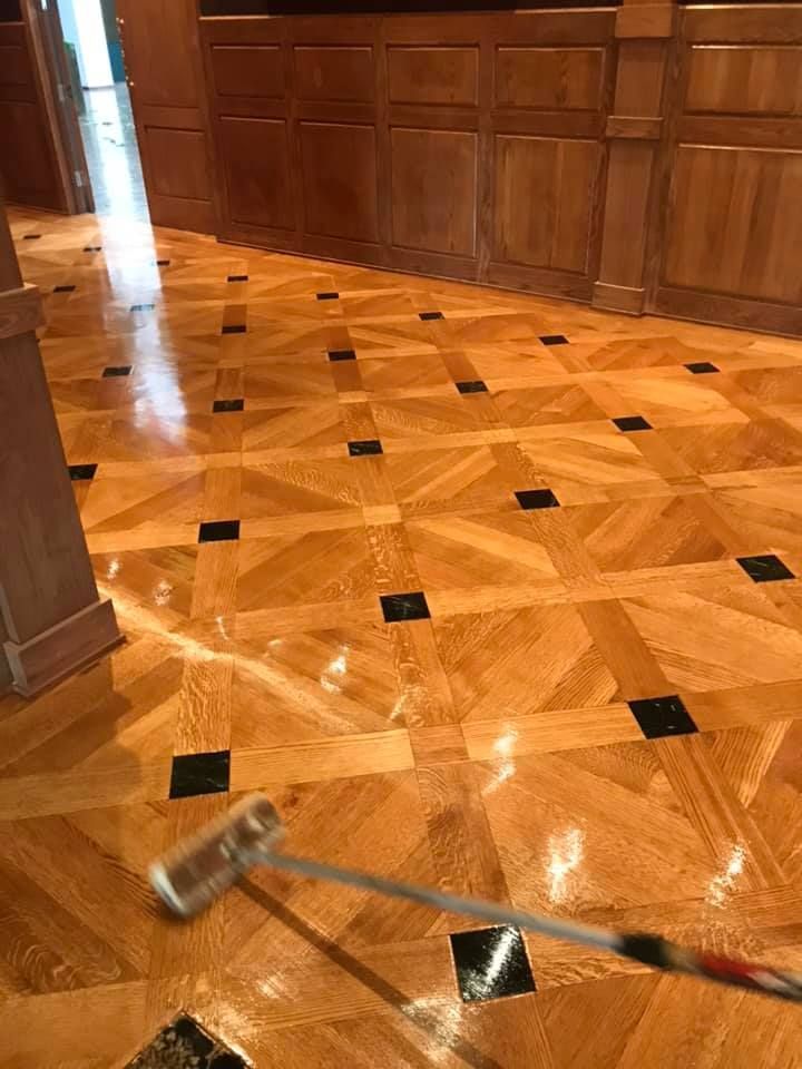 A person applying a glossy finish to a parquet wood floor with black square inlays using a roller.