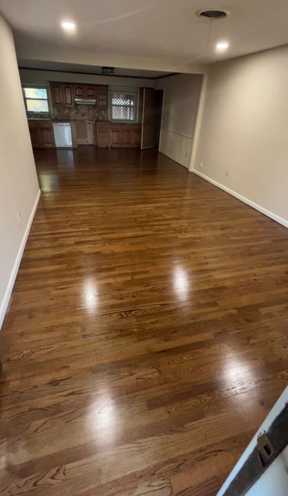 Spacious living area with dark wood floors. Kitchen visible in the background. Bright lighting, neutral walls.