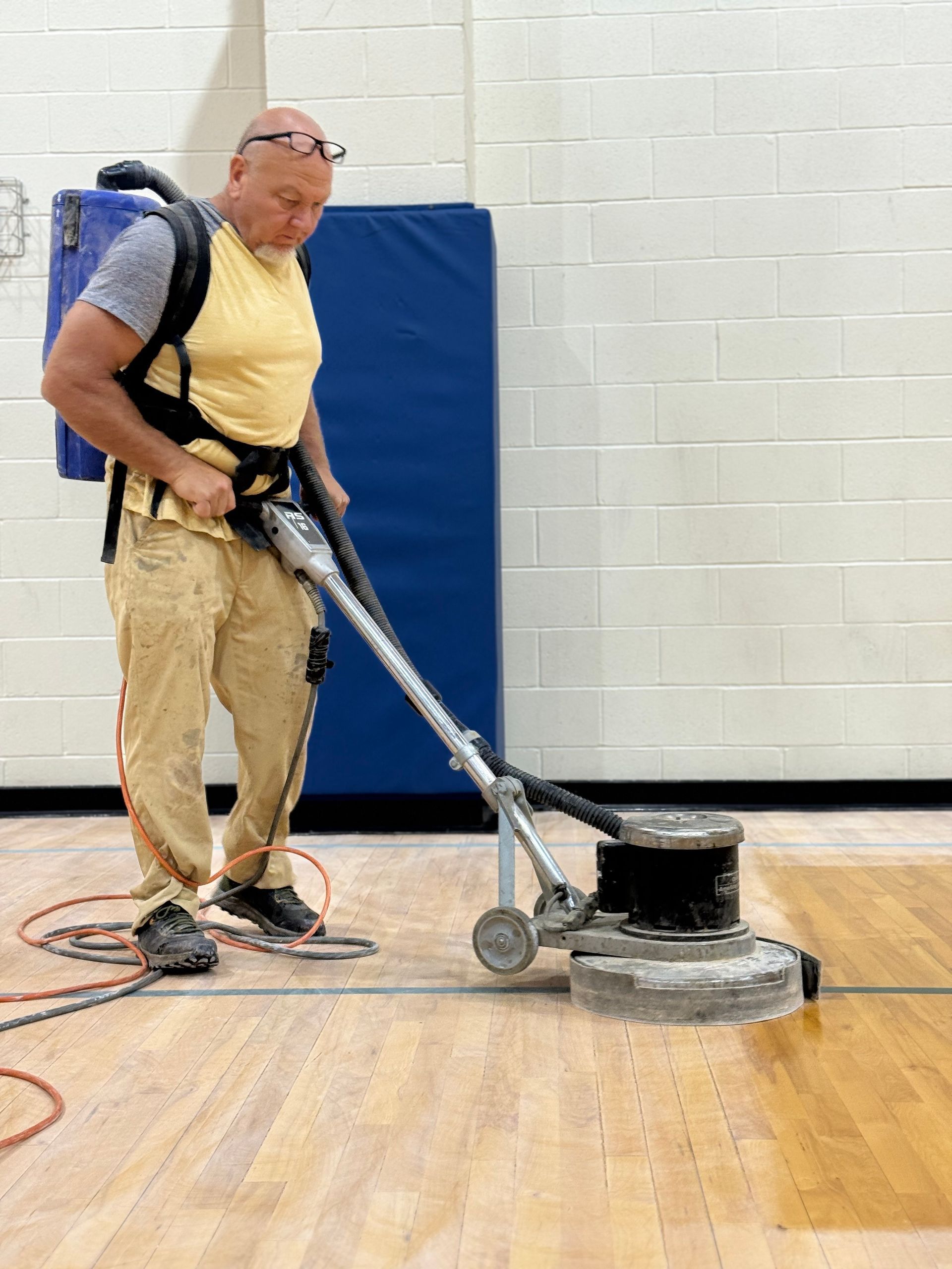 A person sanding a wooden floor in a gymnasium. They are wearing safety glasses and a backpack.