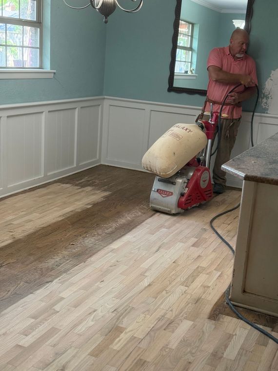 Man sanding hardwood floor in room with wainscoting and blue wall. Dust and sander are visible.