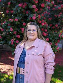 A woman in a pink jacket and glasses is standing in front of a bush of pink flowers.