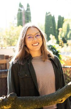 A woman wearing glasses is holding a tree branch and smiling.