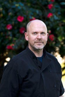 A bald man in a black chef 's jacket is standing in front of a bush with red flowers.