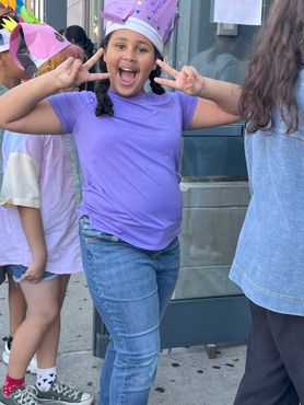 A young girl wearing a purple shirt and a crown giving a peace sign