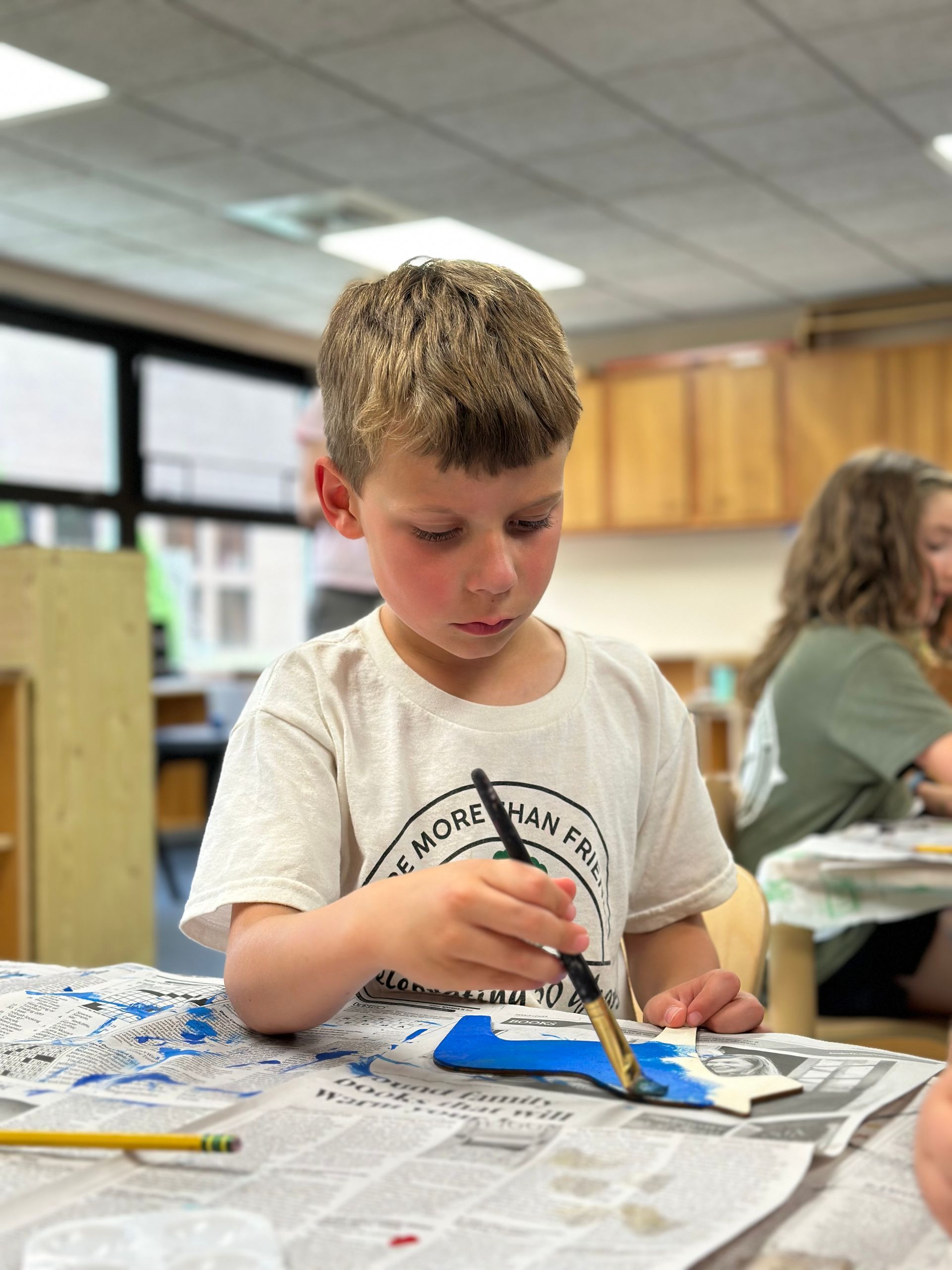 Child painting at a table in a classroom, focused on a blue craft project.