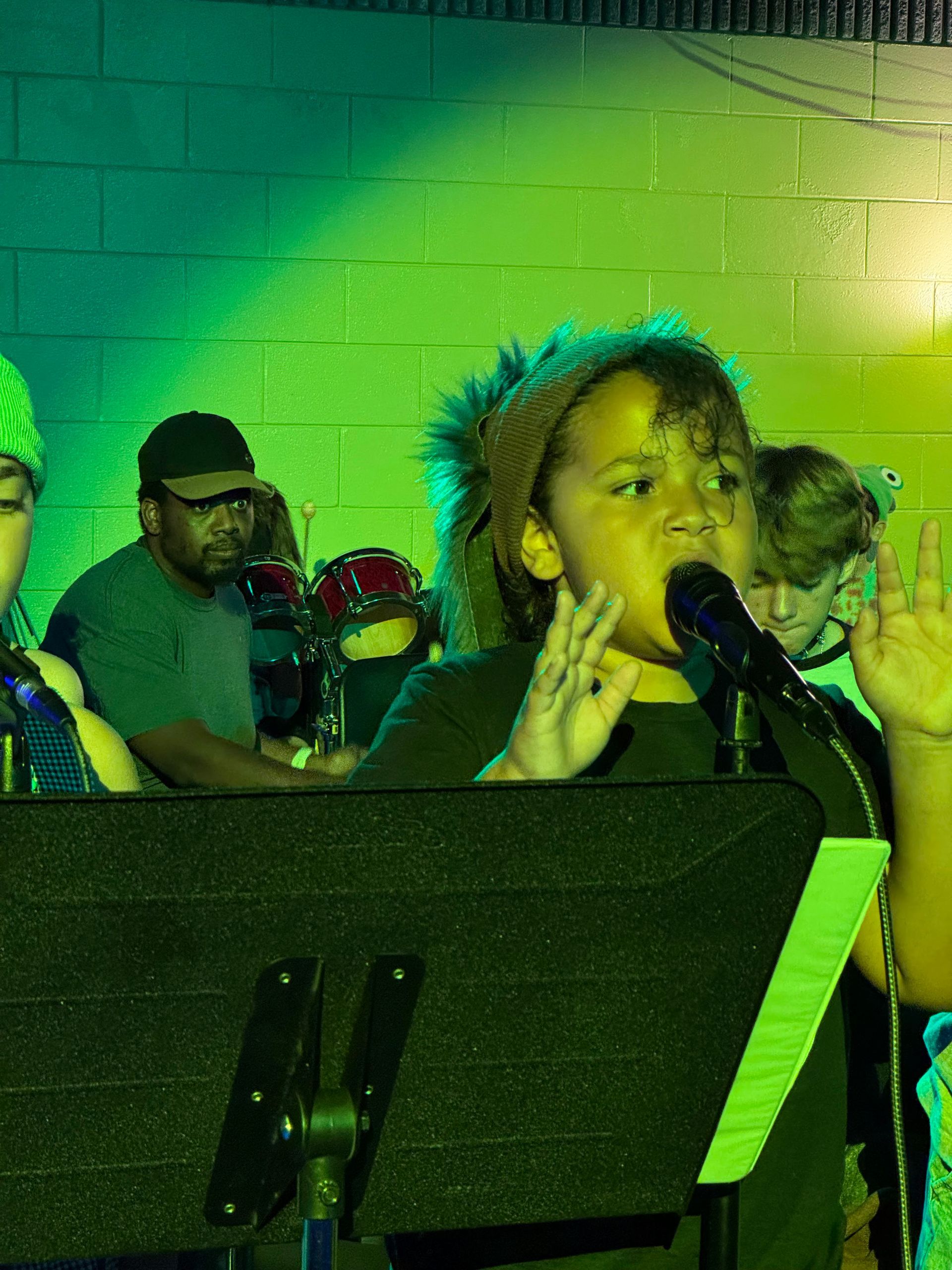 Singer performing into a microphone on a green-lit stage, with a music stand in front and band members behind.