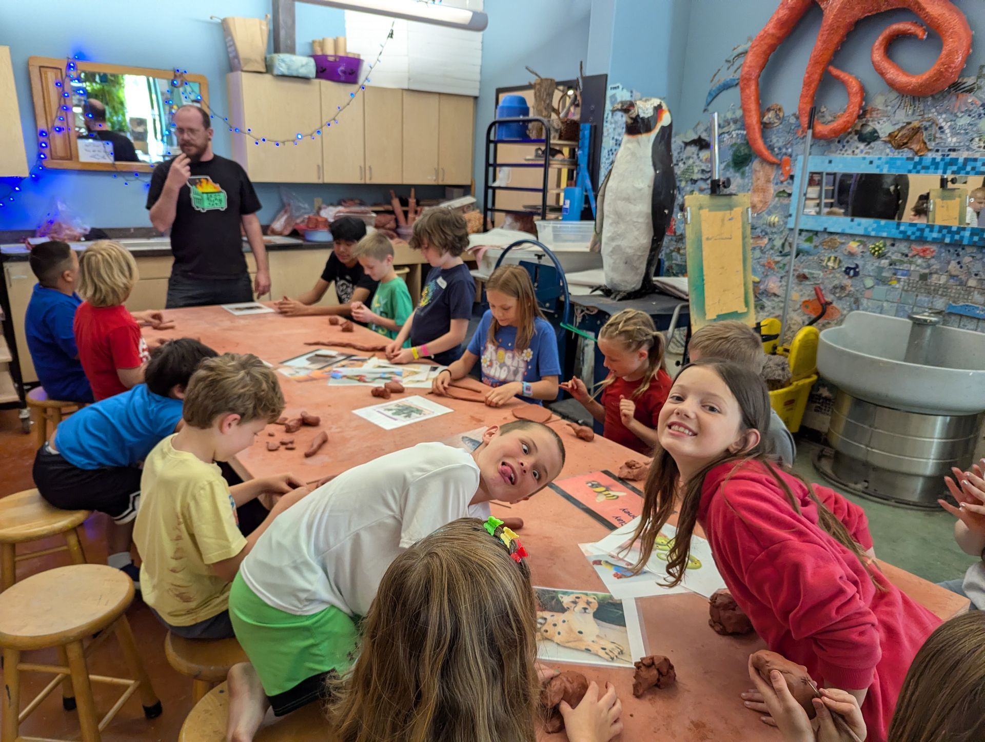 Children painting at a classroom table with a teacher nearby, surrounded by colorful art supplies.