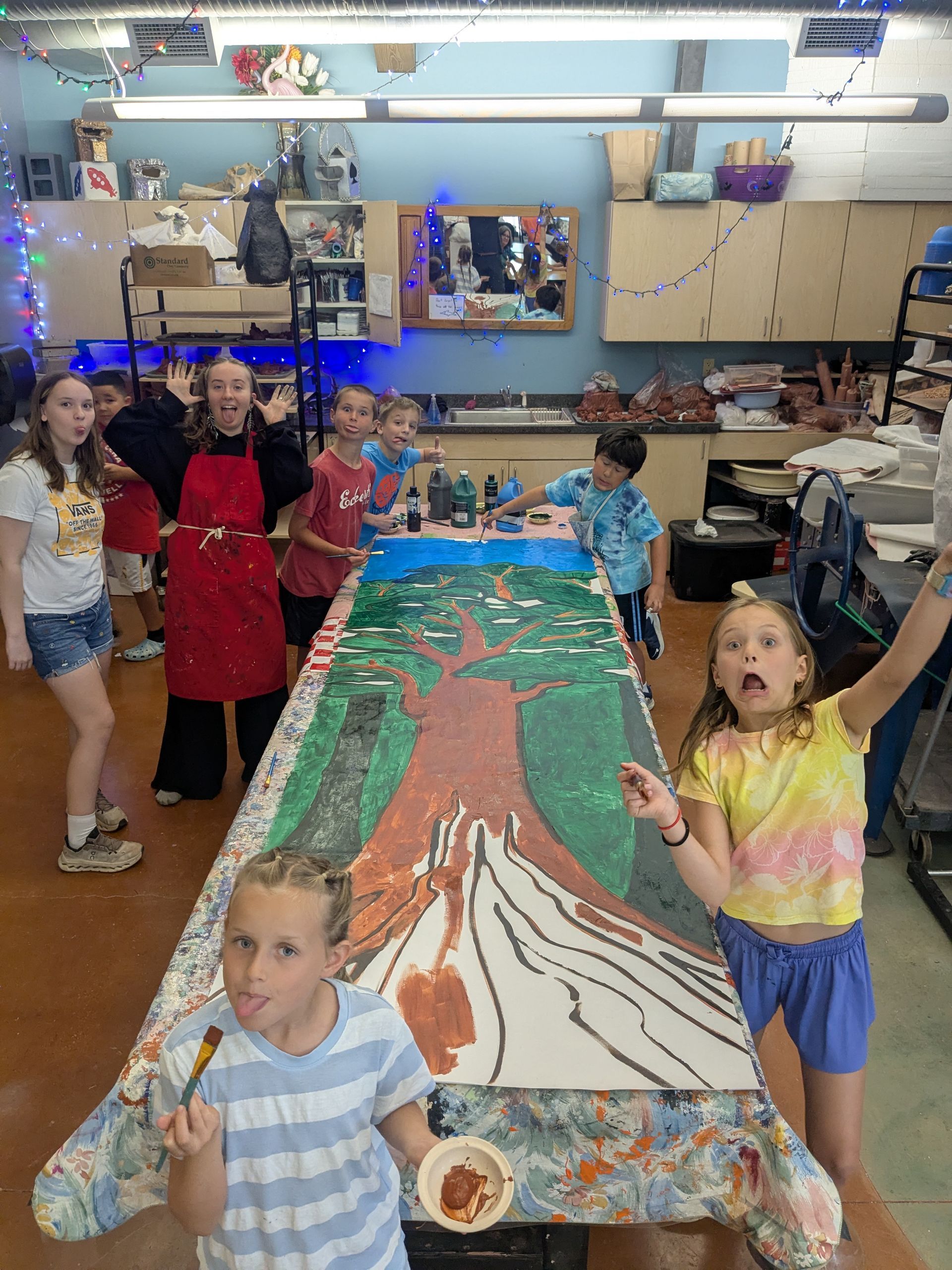 Children in an art classroom surround a long painted table, with one child holding a paintbrush in the foreground.