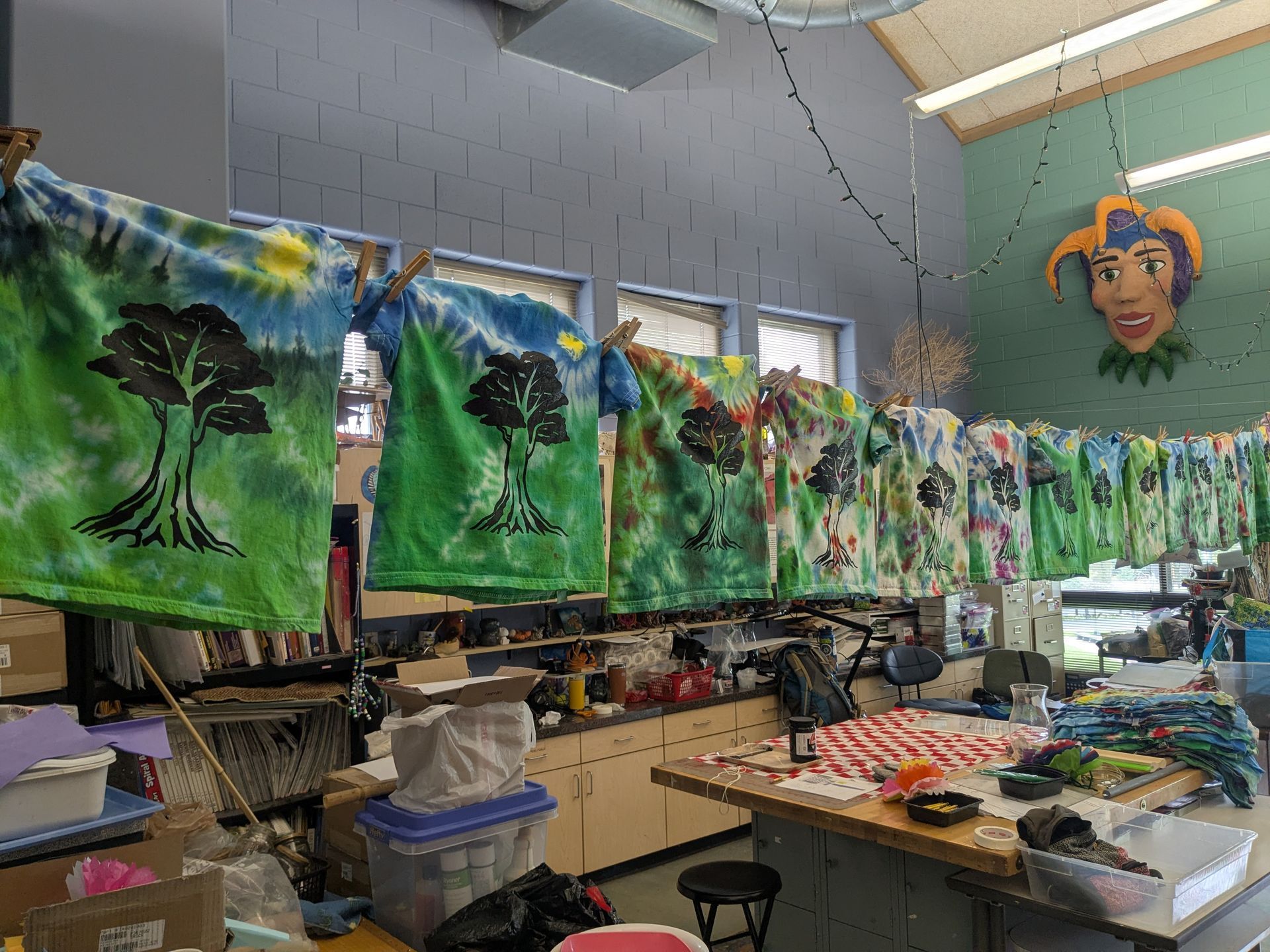Art classroom with hanging painted landscape panels drying above cluttered tables and supplies