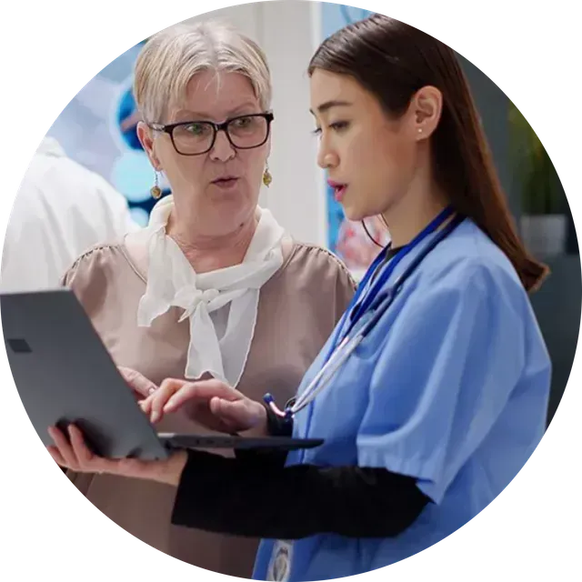 Two healthcare workers reviewing a laptop in a clinic, one in blue scrubs with a stethoscope
