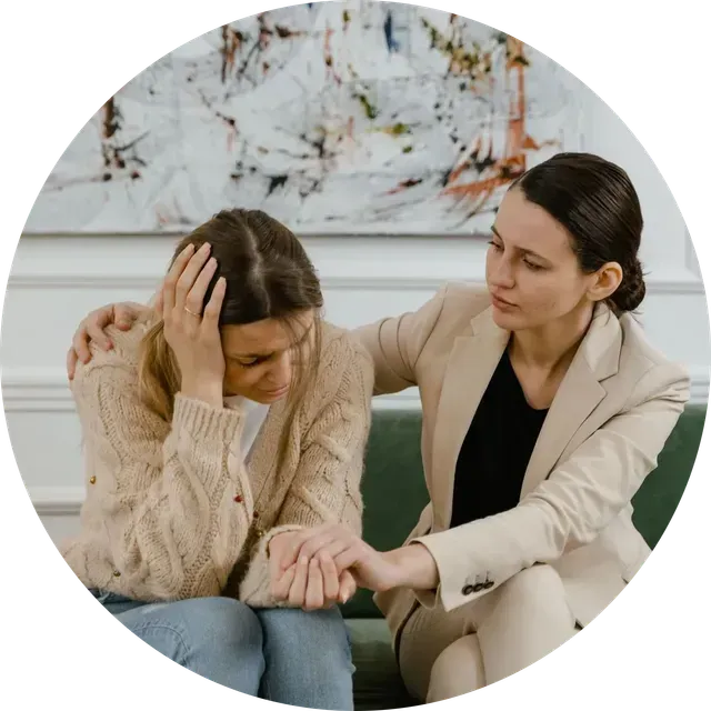 Two people sit on a couch, one comforting the other as she looks distressed.