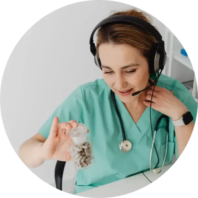 Healthcare worker in teal scrubs wearing a headset, holding a pill bottle and smiling at a phone call