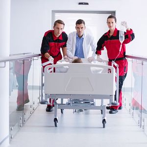 Three paramedics in red uniforms push a hospital bed down a bright hallway.