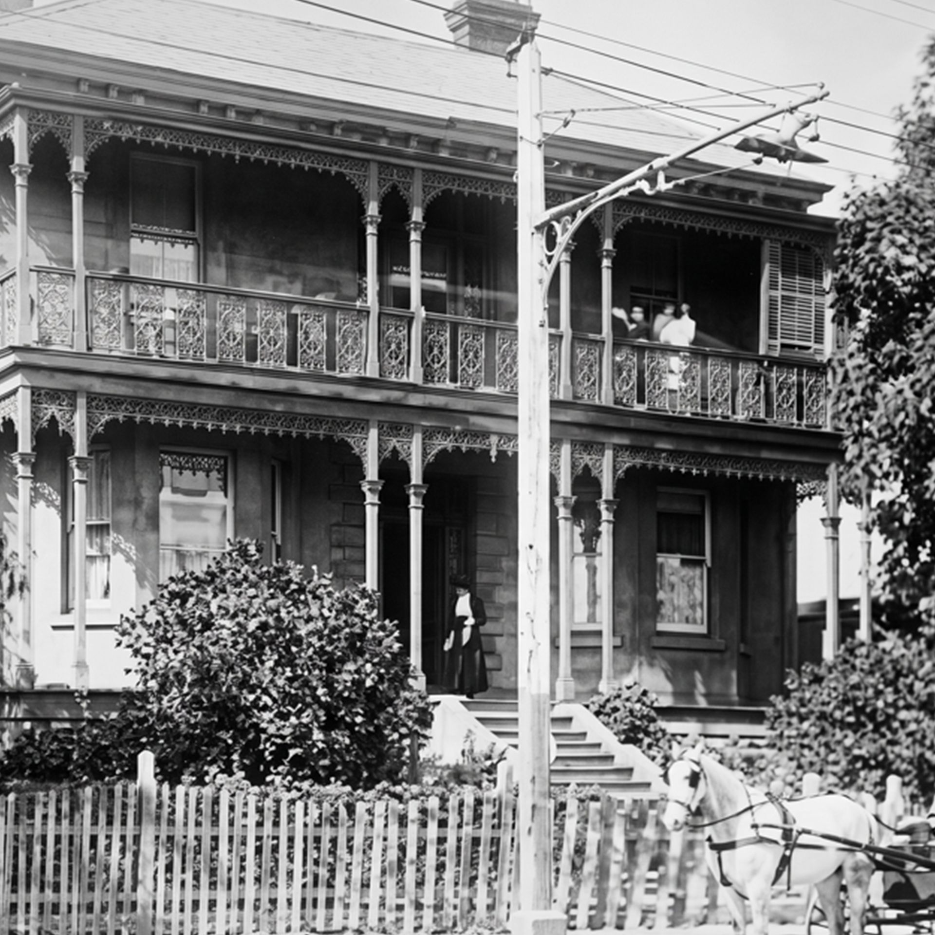 Black-and-white photo of a Victorian house with ornate balconies, picket fence, and horse-drawn carriage in front