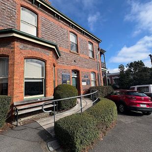 Brick building with ramp and handrail beside trimmed hedges and a red car under a blue sky.