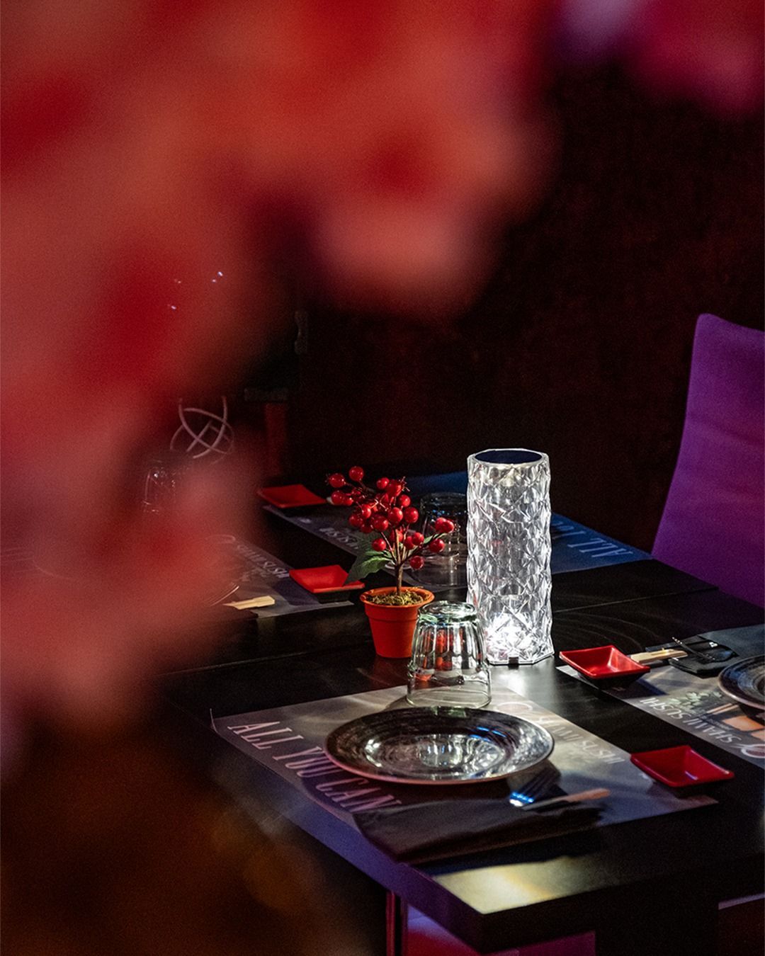 Tavolo del ristorante apparecchiato per la cena: piano nero, lampada in vetro trasparente, decorazione floreale rossa, sedia viola.
