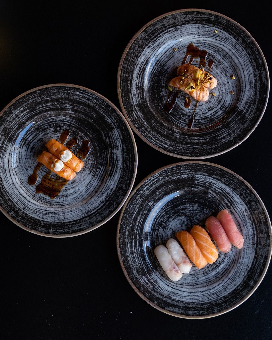 Tre piatti di sushi su una superficie nera. Ognuno contiene pezzi diversi, tra cui salmone e tonno, con salse e condimenti.