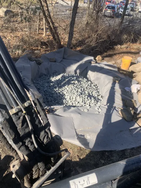 Pile of gray gravel on a tarp beside black pipes and a yellow bucket in an outdoor yard.