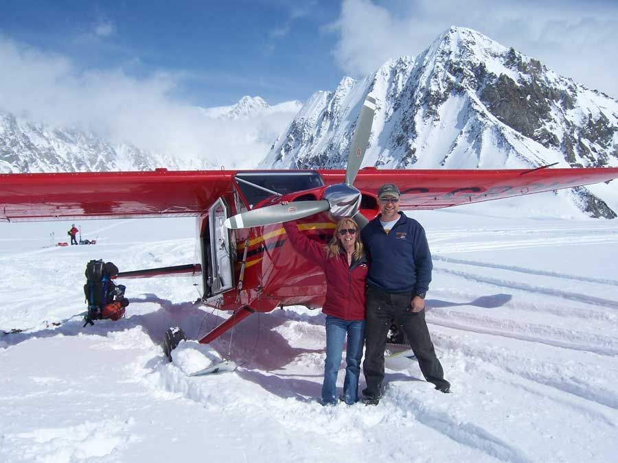 Two people beside a red airplane on snowy mountains under a bright sky