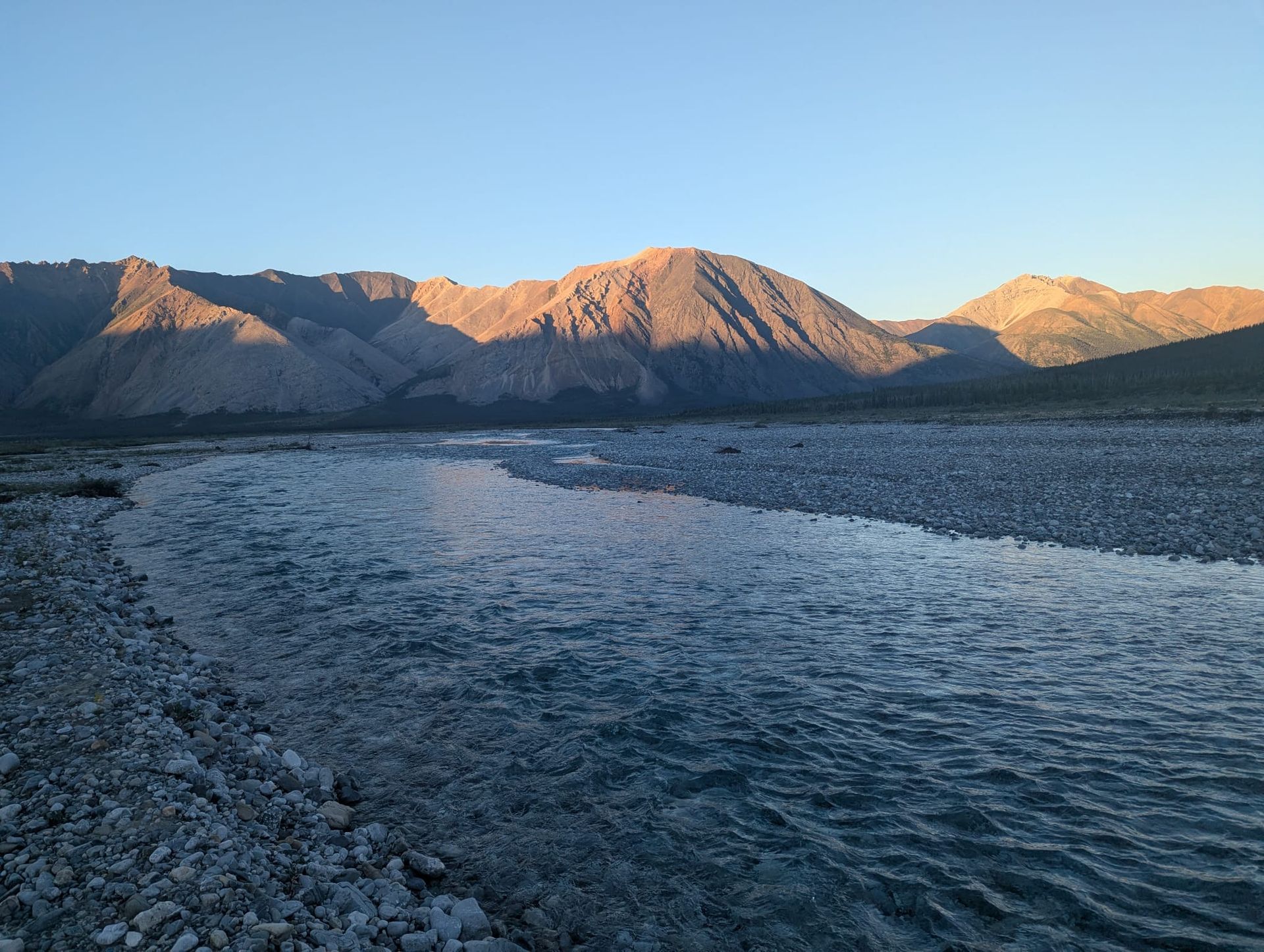 Rocky riverbank at sunset with blue water and sunlit mountains in the distance