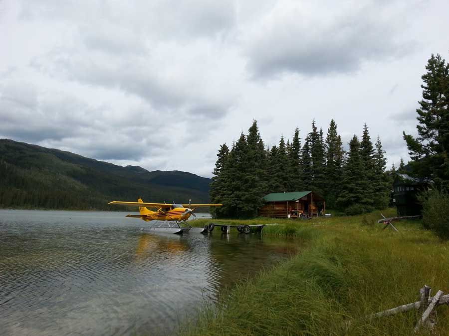 Yellow floatplane on a lake beside a cabin and pine trees under cloudy mountains