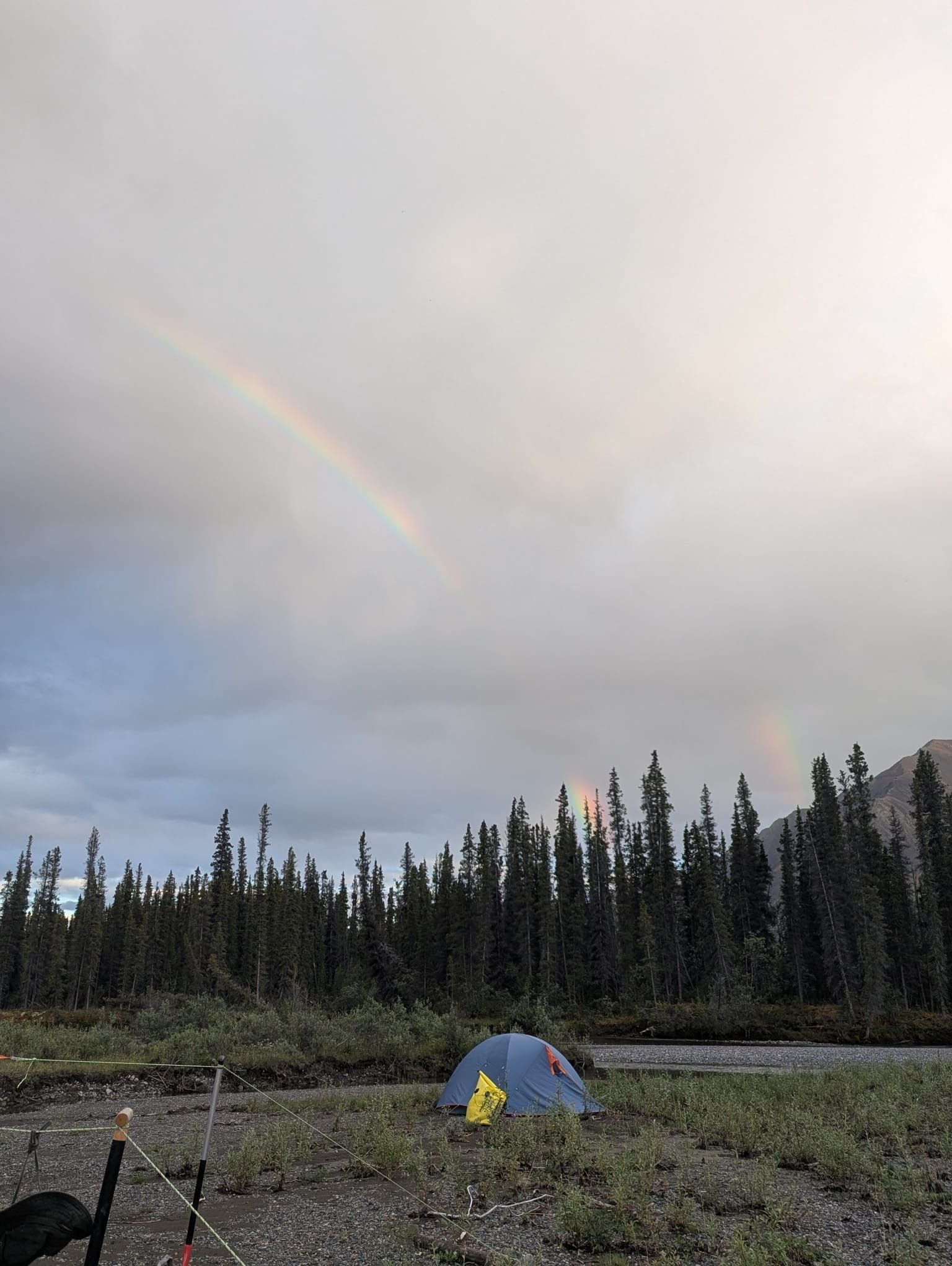 Tent on a rocky field beneath a faint rainbow and cloudy sky, with a dark evergreen forest in the background