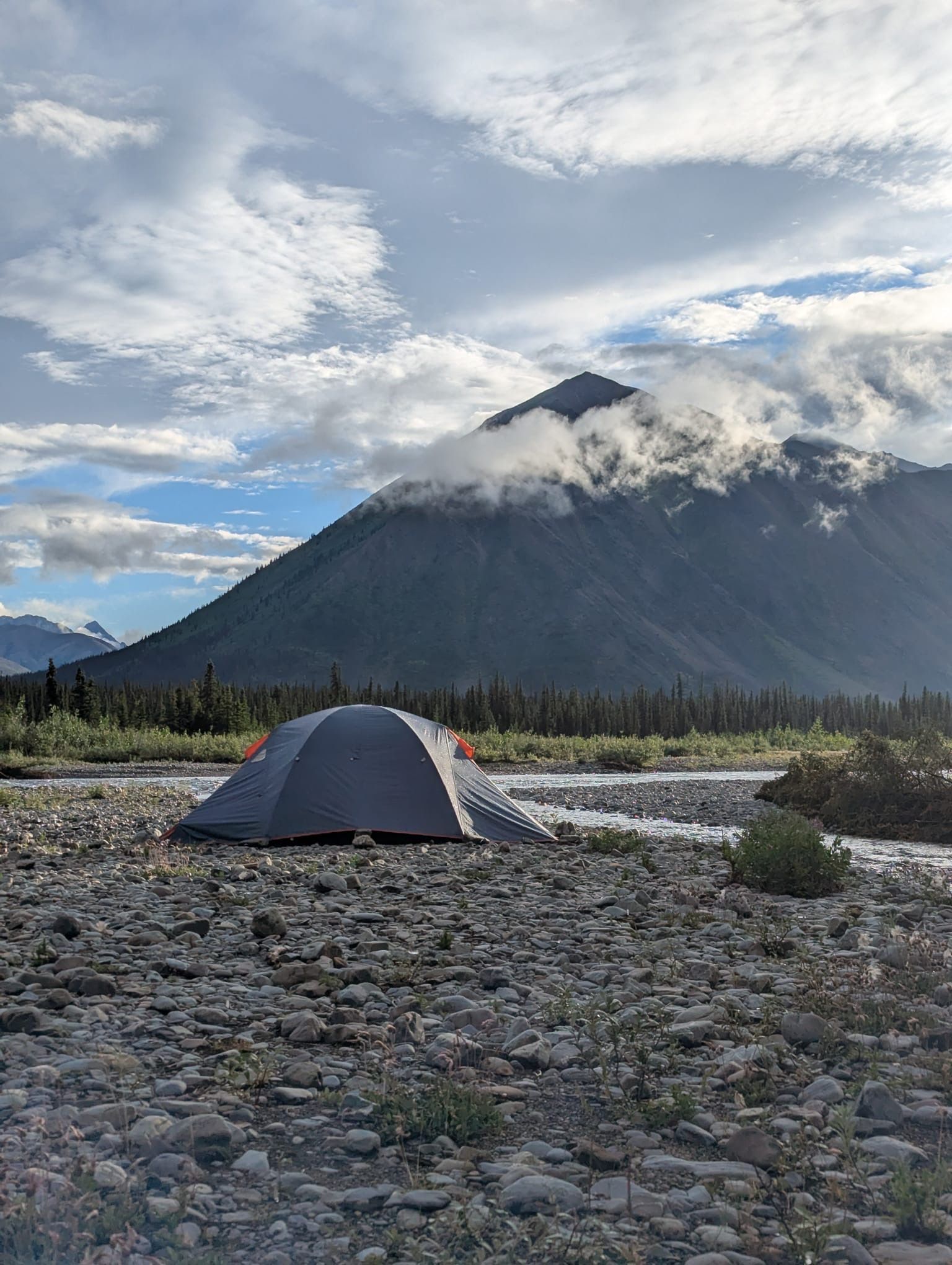 Tent pitched on rocky ground beneath a cloud-wrapped mountain under a partly cloudy sky