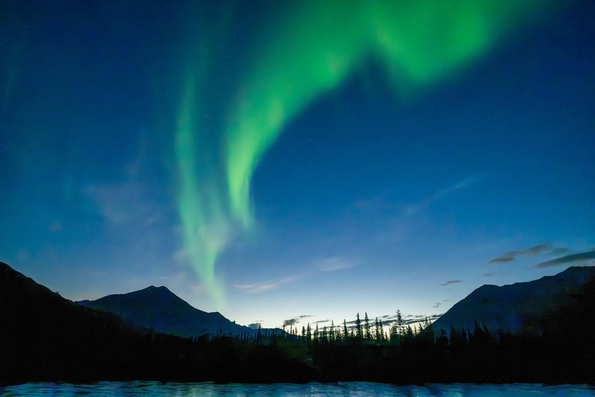 Green aurora over dark mountain silhouettes and a forest at twilight