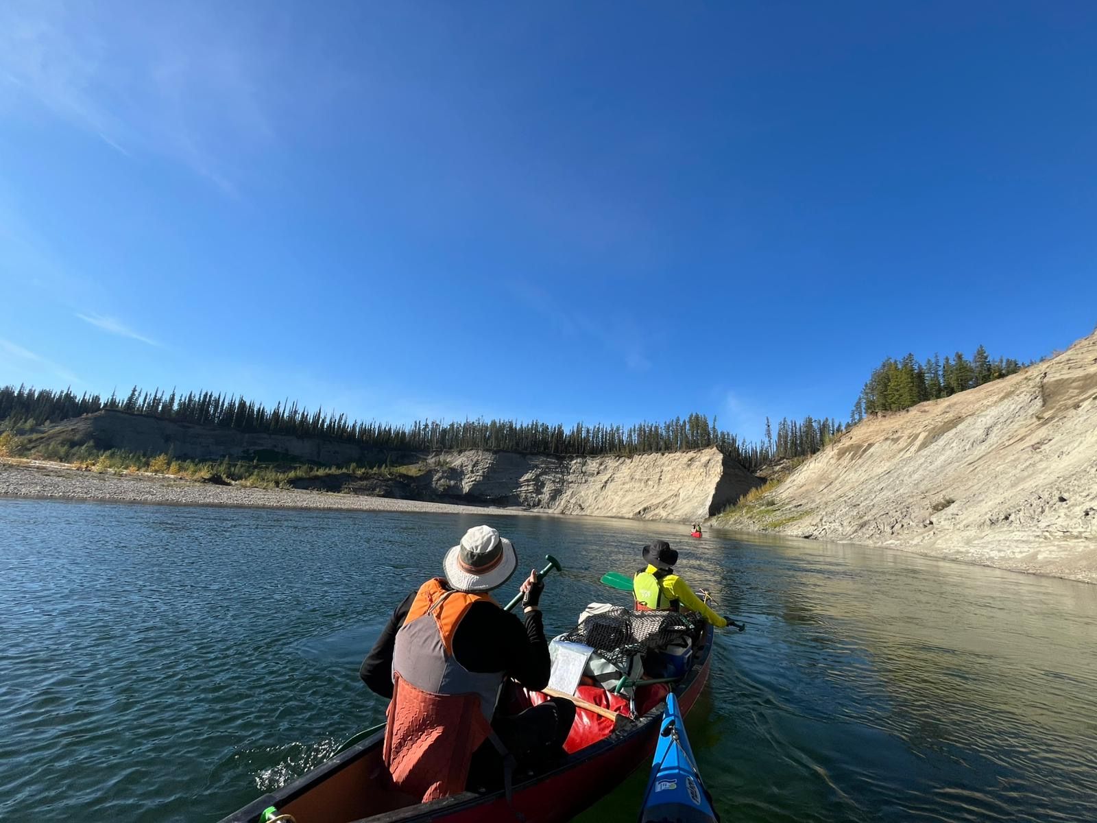 People in kayaks on a calm blue river beside steep sandy banks under a clear sky
